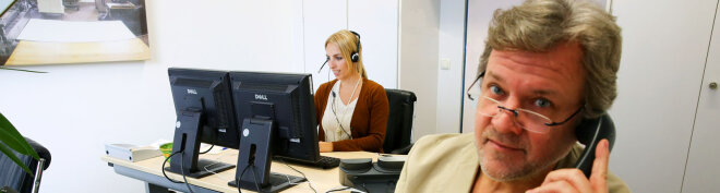 eine Frau an einem Bildschirmarbeitsplatz, rechts ein Mann mit Telefonhörer